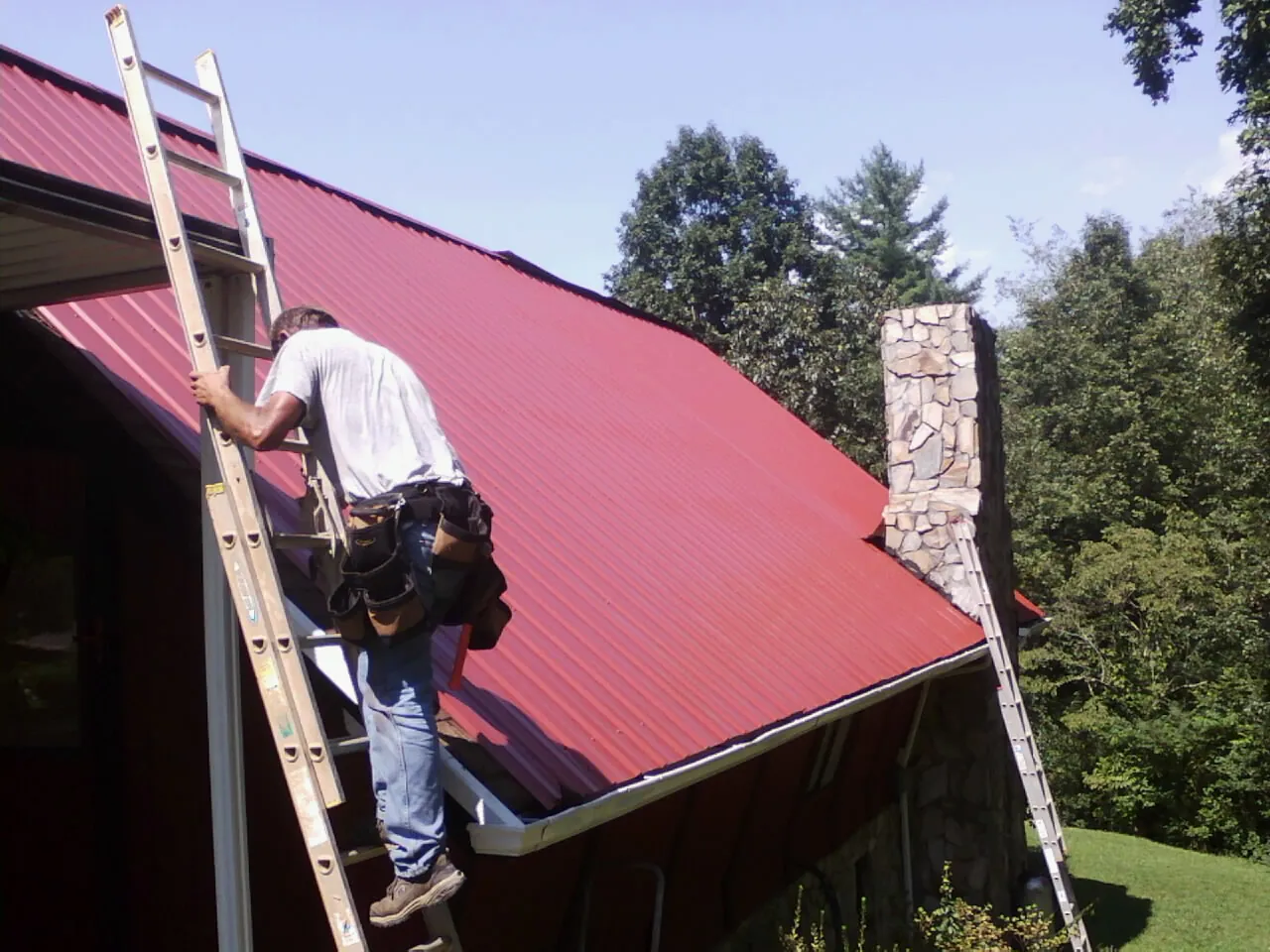 Roofer on a ladder beside a red metal roof and stone chimney — Soffit & Fascia Repair by Roofing Brothers in Madison